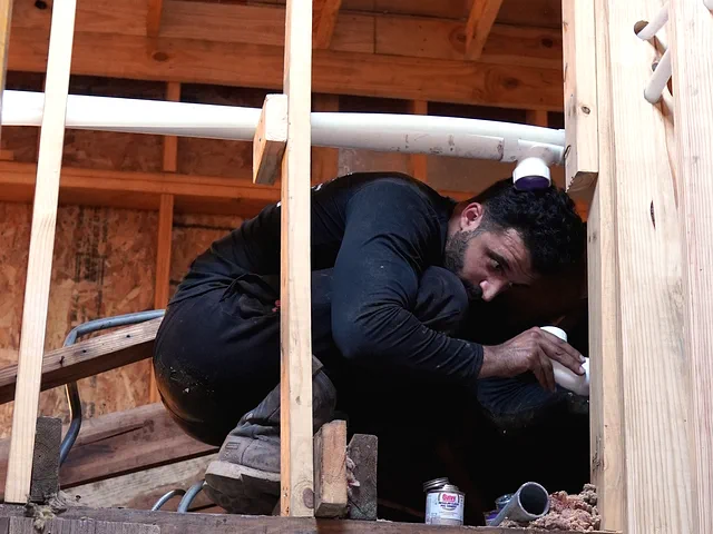 A crew member crouched up in the roof framing, checking a white PVC fitting under an overhead pipe run.