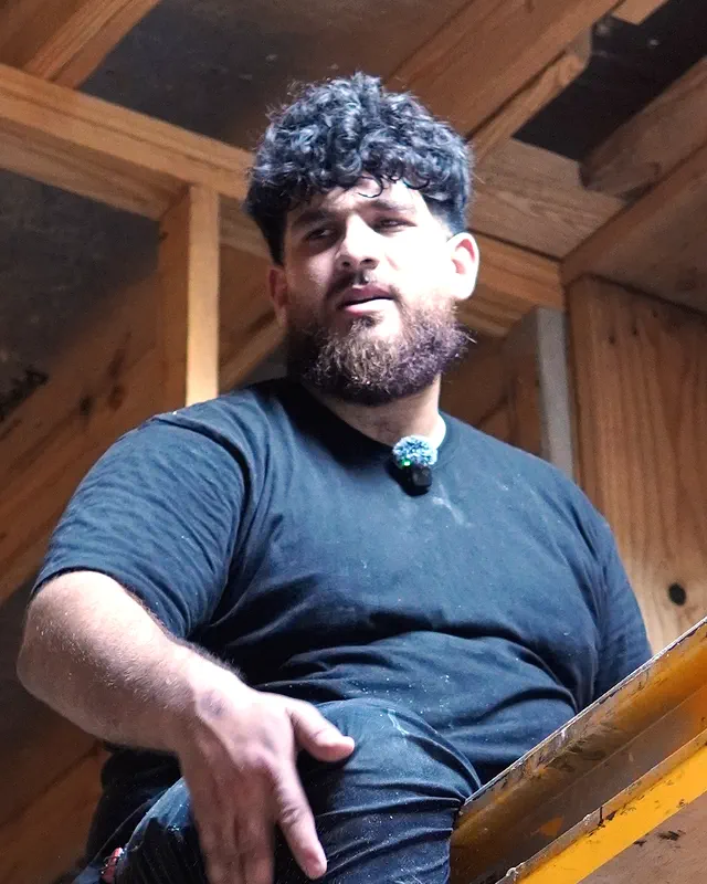 The owner of H&S Plumbing in a black T-shirt, looking down at the camera from a scaffold under the roof framing.