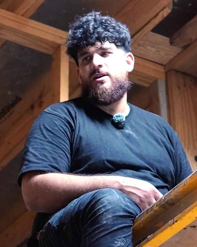 The owner of H&S Plumbing sitting on a scaffold under the roof framing, talking.